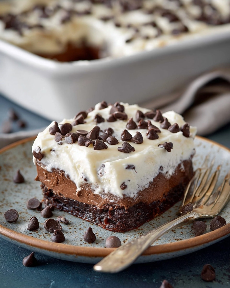 The image showcases a decadent slice of chocolate dessert topped with a generous layer of creamy frosting sprinkled with mini chocolate chips. The dessert has multiple layers, including a chocolate base and a rich, smooth filling beneath the frosting, creating a visually appealing contrast of textures and flavors. The slice is served on a rustic, speckled plate with a vintage-style fork, with additional chocolate chips scattered around, enhancing the overall indulgent presentation. In the background, the remaining dessert in a white baking dish is slightly blurred, keeping the focus on the slice in the foreground. photo taken with an iPhone --ar 4:5 --v 7