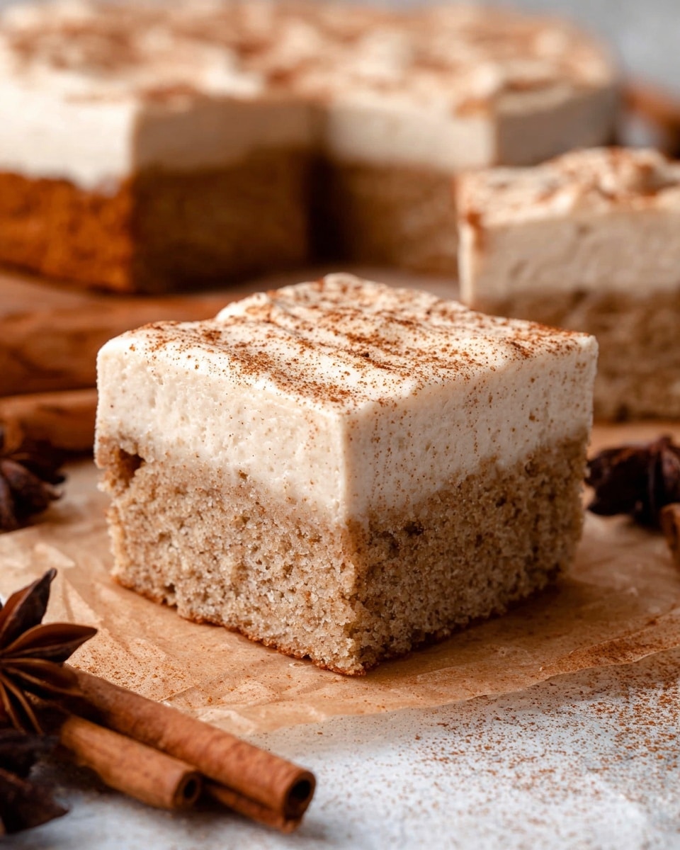 A close-up view of a single square piece of cake with two layers resting on parchment paper over a wooden surface. The bottom layer is dense and light brown with a coarse, crumbly texture. The top layer is thick, creamy, and pale beige with a smooth finish, sprinkled lightly with brown spice powder. In the background, there is a round cake with similar layers, partially cut into squares, and some whole cinnamon sticks and star anise are placed nearby. The surface is white marbled texture. photo taken with an iphone --ar 4:5 --v 7