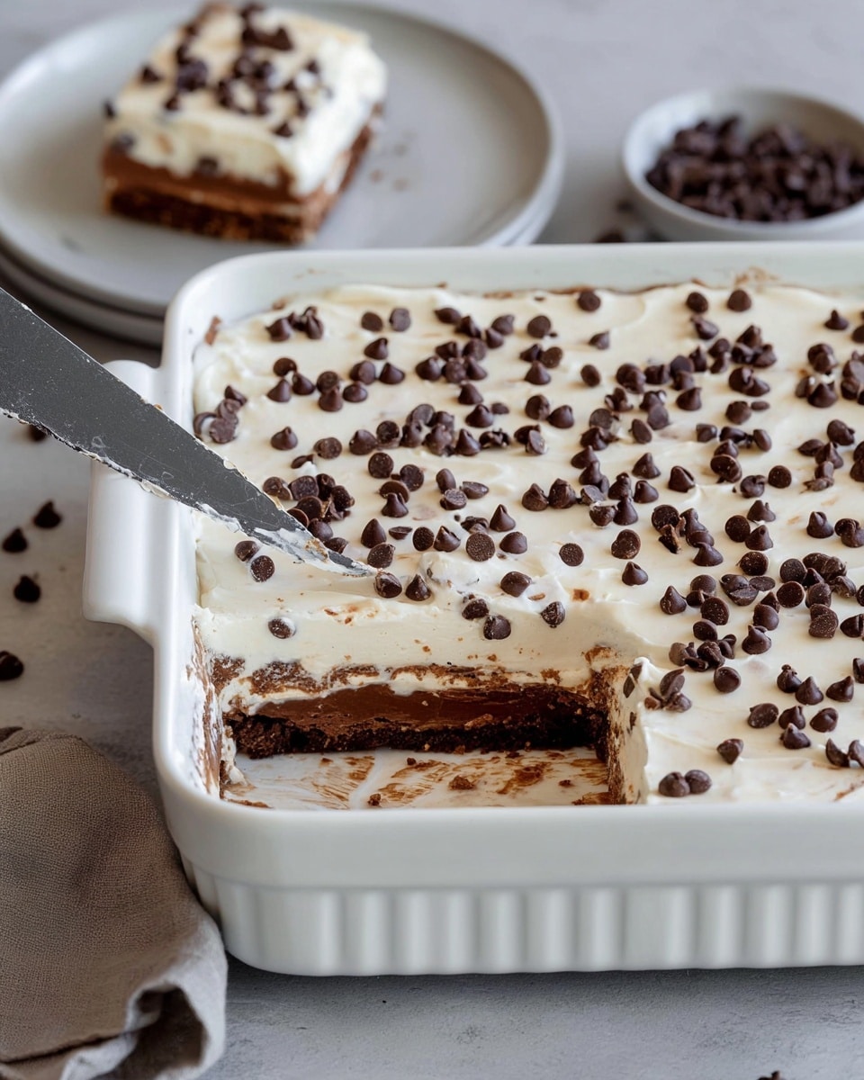 This image showcases a rich layered dessert in a white ceramic baking dish. The bottom layer appears to be a dense chocolate base, topped with a thick, creamy layer generously sprinkled with mini chocolate chips. A knife is cutting a square portion of the dessert, revealing the distinct chocolate layer beneath the creamy topping. In the background, a piece of the sliced dessert is placed on a small plate, ready to be enjoyed. Photo taken with an iPhone --ar 4:5 --v 7