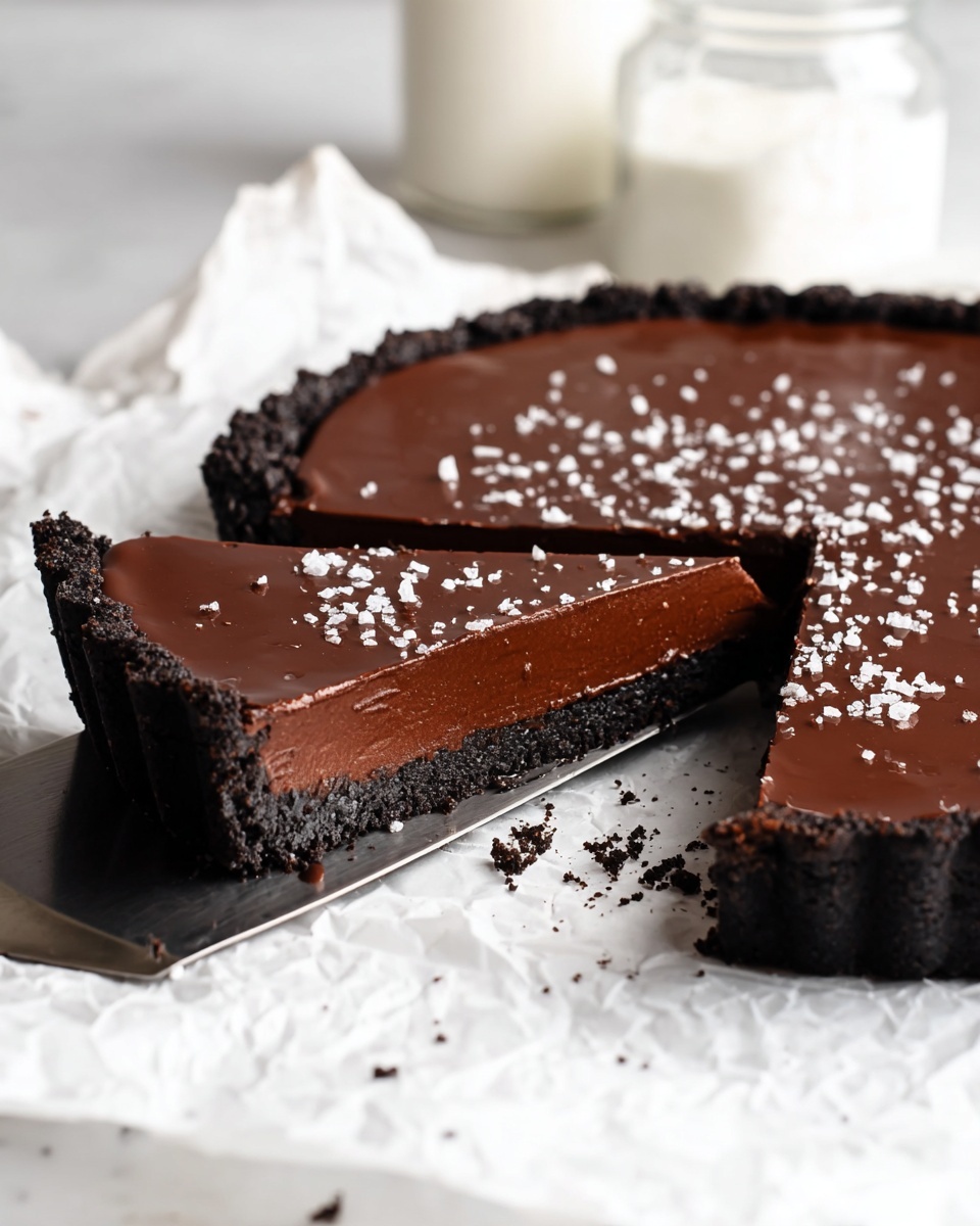 A dark chocolate tart is shown with one large slice cut out and slightly pulled forward on a metal spatula. The tart has three visible layers: a black crumbly crust on the bottom and sides, a thick middle layer of smooth, dark chocolate filling with a rich texture, and a glossy dark chocolate ganache on top, sprinkled with small white salt flakes. The tart rests on crumpled white paper placed on a white marbled surface. In the soft background, there is a blurry glass jar with white cream inside. photo taken with an iphone --ar 4:5 --v 7