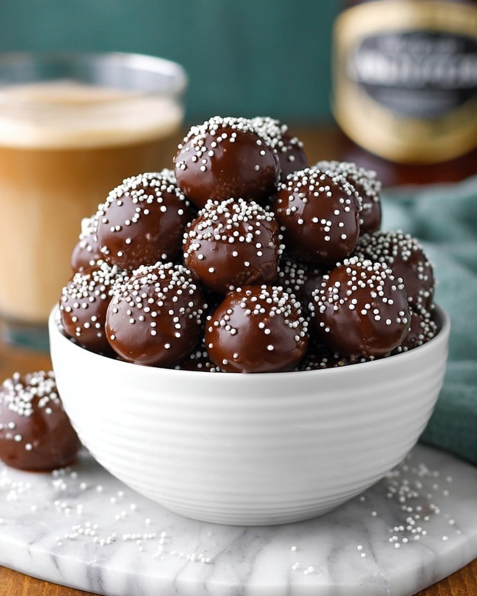 A white bowl filled with many round chocolate balls covered in smooth, shiny dark brown chocolate, each sprinkled with tiny white round sprinkles. The balls are stacked in layers, rising above the bowl’s rim, showing a rich, glossy texture. The bowl sits on a white marbled surface with a few scattered white sprinkles around it. In the background, a glass with a layered creamy drink and a blurred bottle can be seen. Photo taken with an iphone --ar 4:5 --v 7