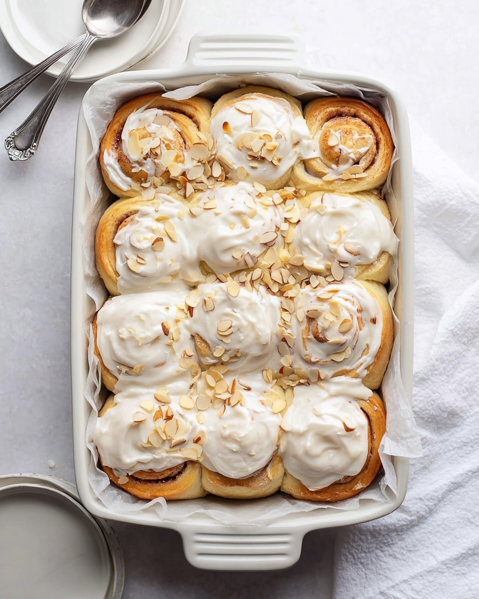 A white baking dish holds eight soft, fluffy cinnamon rolls, with one roll missing from the front left corner, revealing the light, airy texture inside. Each roll is topped with a creamy white icing that spreads unevenly over the surface, and pale, thinly sliced almond pieces are scattered generously on top, adding a slight crunch. The rolls are set against a white marbled surface, and the baking dish is lined with parchment paper peeking out slightly around the edges. photo taken with an iphone --ar 4:5 --v 7