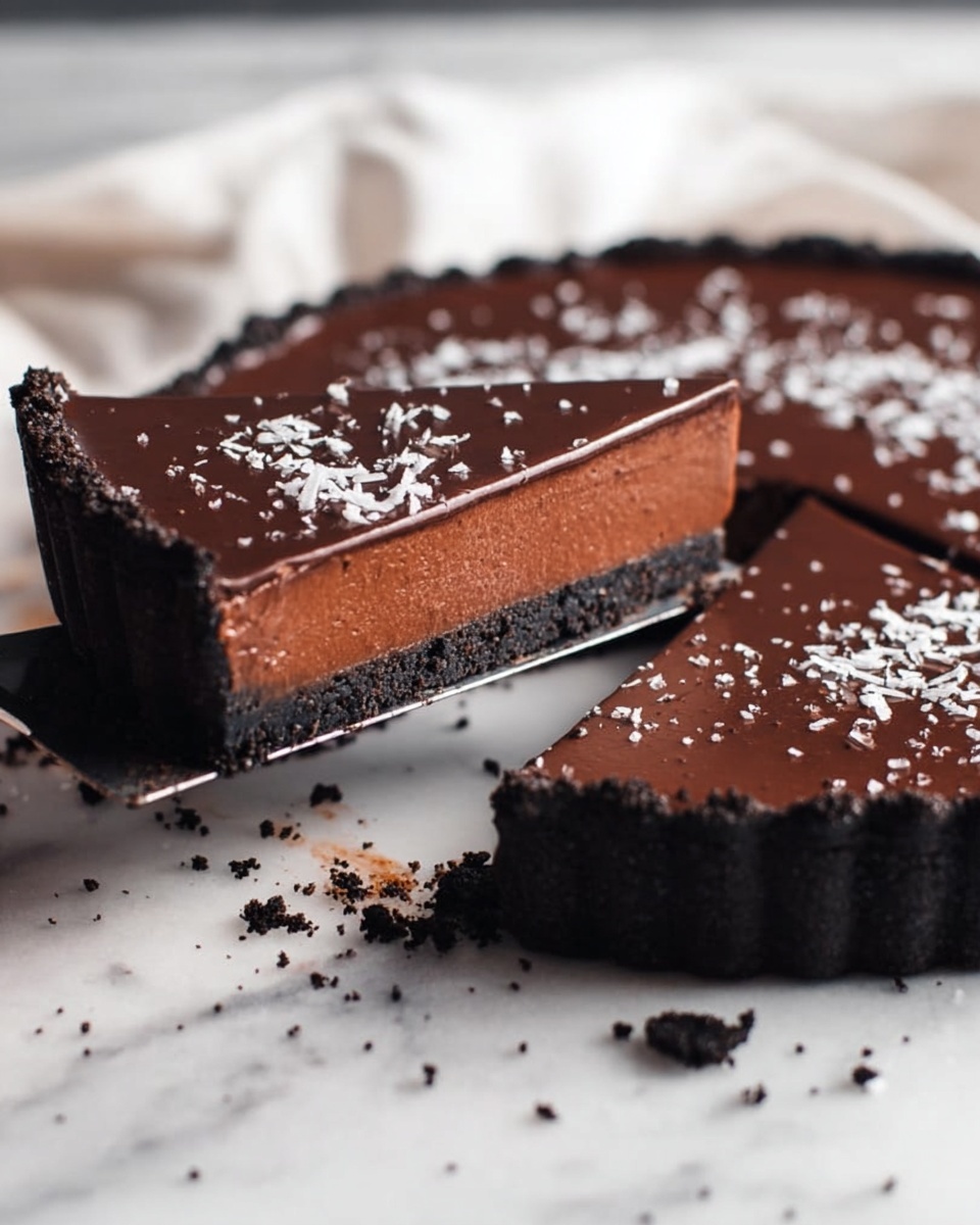 The image shows a chocolate tart with three visible layers on a white marbled surface. The bottom layer is a dark, crumbly crust made of crushed cookies, forming a thick edge around the tart. The middle layer is a rich, smooth chocolate filling that looks creamy and dense, with a lighter brown color than the top. The top layer is a shiny, darker chocolate glaze, sprinkled with white flakes that add texture and contrast. One slice is lifted on a metal spatula, with a woman's hand holding the spatula just out of frame. Crumbs are scattered around the tart on the white marbled surface. Photo taken with an iphone --ar 4:5 --v 7