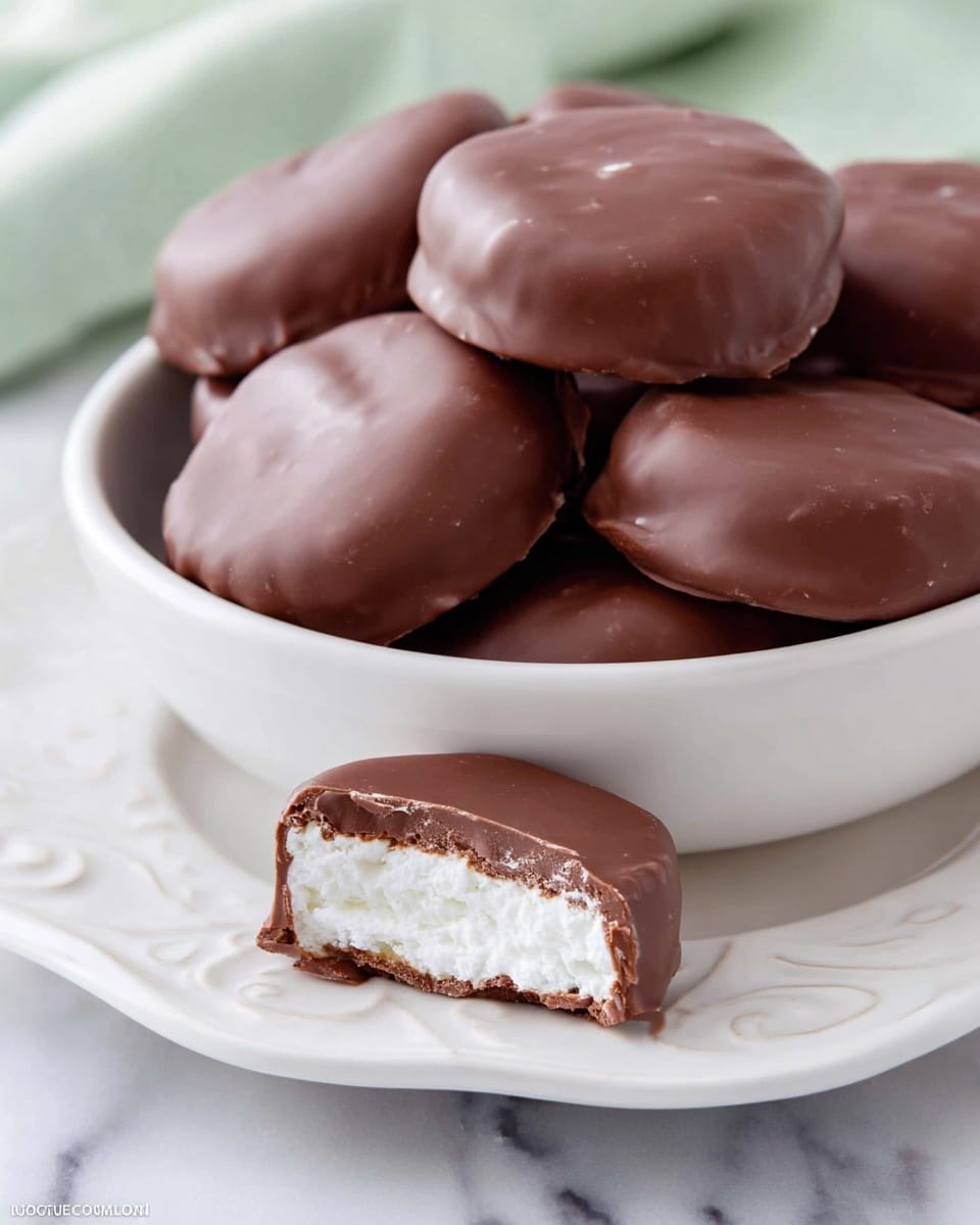 A white bowl is filled with round chocolate-covered treats, each smooth and shiny with a milk chocolate coating. The treats are stacked, showing their dark brown chocolate outer layer, with one treat on top cut open to reveal a white, creamy, and slightly textured filling inside. The bowl is sitting on a green plate with a raised pattern around the edges. The scene is set on a white marbled surface. photo taken with an iphone --ar 4:5 --v 7