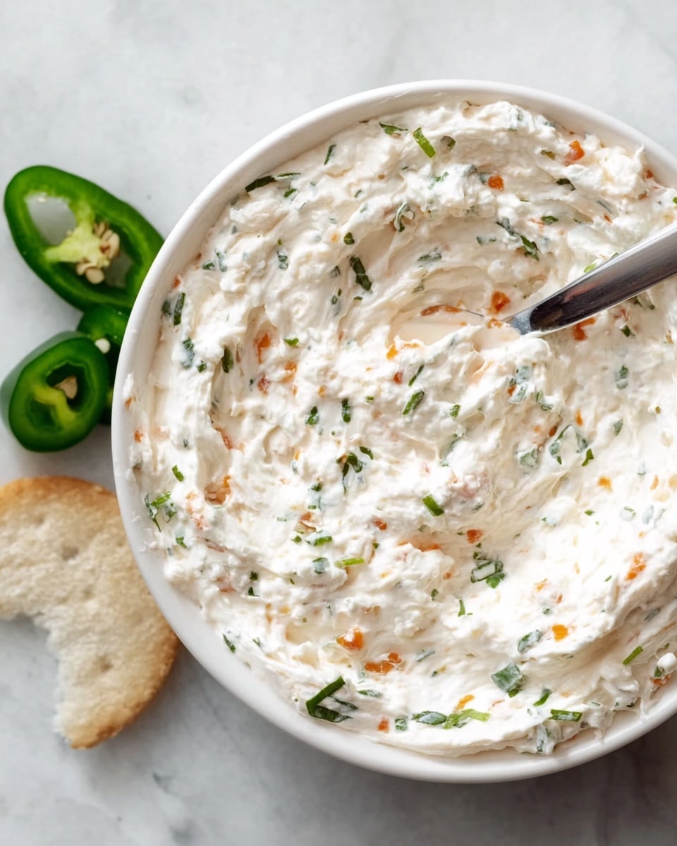 A close-up of a creamy, white dip in a shallow white bowl on a white marbled surface. The dip has a soft, smooth texture mixed with small pieces of green herbs and tiny orange bits scattered throughout. A silver spoon is partially dipped into the mixture, creating gentle swirls on the surface of the dip. Two slices of green jalapeño pepper are placed nearby on the white marbled background. photo taken with an iphone --ar 4:5 --v 7