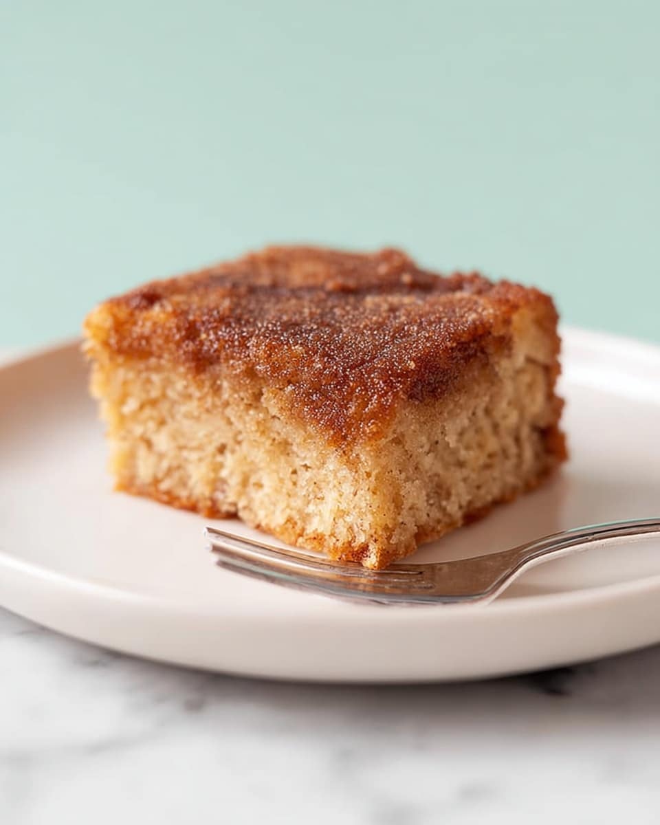 A single square piece of cinnamon cake sits on a white plate, showing two layers: a thick, light brown, moist and crumbly base with a slightly rough texture, and a darker brown, sugary, cinnamon topping with a grainy texture that looks slightly crunchy. The cake is positioned toward the right side of the plate, and a small silver fork rests on the left edge of the plate, all set against a white marbled texture background. photo taken with an iphone --ar 4:5 --v 7