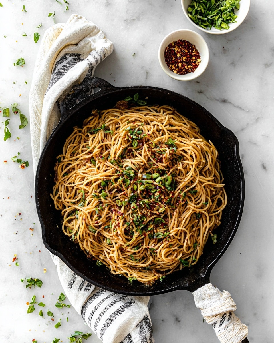 A black cast iron pan holds a single layer of cooked noodles mixed with a light brown sauce. The noodles are thin and twisted together, topped with small green chopped herbs and red chili flakes scattered unevenly over them. The pan’s handle is wrapped with a white and black striped cloth. Above the pan, two small white bowls contain chopped green herbs and red chili flakes. The whole scene sits on a white marbled surface with some scattered green herbs. photo taken with an iphone --ar 4:5 --v 7