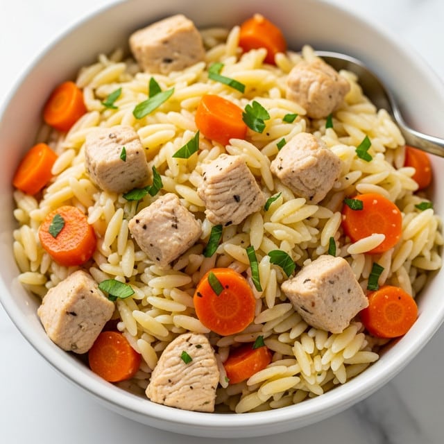 A close-up view of a bowl filled with a creamy orzo dish showing three main layers: small orzo pasta pieces in an off-white color form the base, mixed evenly with tender, light brown chicken cubes scattered throughout; small bright orange carrot cubes add color and texture, while finely chopped green herbs are sprinkled on top, giving a fresh look. The bowl is white and sits on a white marbled surface, with a spoon partially visible inside the dish. photo taken with an iphone --ar 4:5 --v 7
