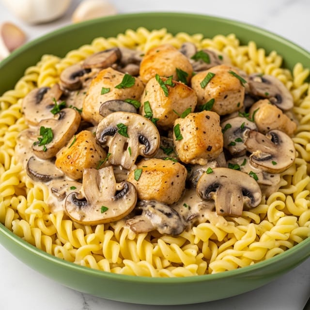 The dish shows a large green bowl filled with twisted egg noodles forming the base layer, pale yellow in color and soft in texture. On top, there is a creamy mushroom sauce layer containing browned mushroom slices and chunks of golden-brown chicken, fully coated in a thick light beige sauce with specks of herbs. Small pieces of bright green parsley are scattered over the top, adding color and freshness. The bowl rests on a white marbled surface, with some garlic cloves blurred in the background. photo taken with an iphone --ar 4:5 --v 7