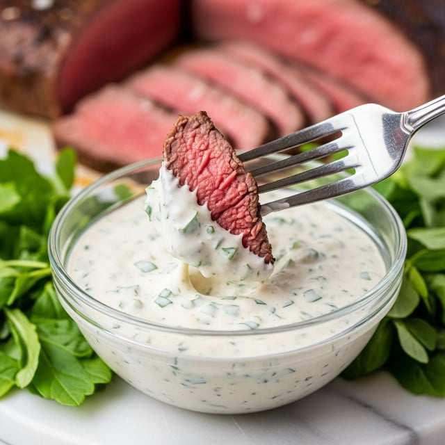 The image shows a close-up of a silver fork holding a piece of medium-rare cooked beef dipped into a thick, creamy white sauce with small green herb bits mixed in. The sauce is in a clear glass bowl placed on a white marbled surface. Fresh green herbs surround the bowl, and more slices of the pinkish-red beef are visible blurred in the background. The focus is on the fork with the beef and sauce, highlighting the creamy texture and tender meat. Photo taken with an iphone --ar 4:5 --v 7
