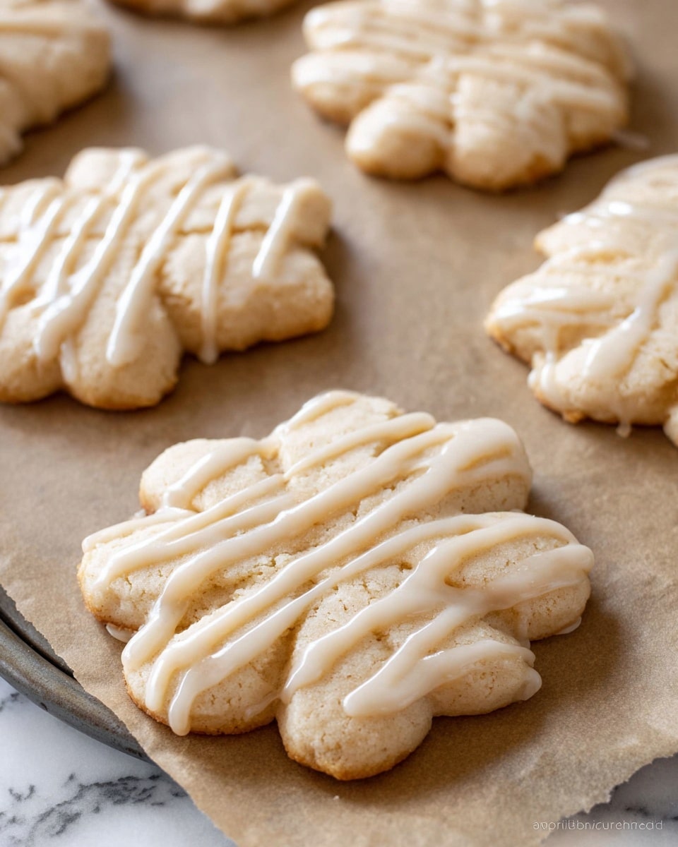 The image shows several pale beige flower-shaped cookies arranged on parchment paper over a baking tray. Each cookie is topped with a smooth, white icing drizzled in thin, uneven lines across the surface. The texture of the cookies looks soft and slightly crumbly, with gently rounded edges and a delicate thickness. The background features a white marbled texture, adding a clean and light setting to the arrangement. photo taken with an iphone --ar 4:5 --v 7