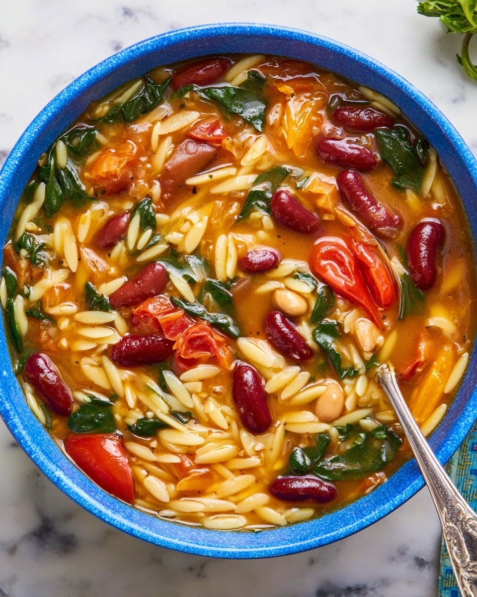 A close-up image of a blue bowl filled with a thick soup showing three main layers: the bottom layer is an orange-tinted broth, the middle layer has small grain-shaped orzo pasta in pale yellow color mixed throughout, and the top layer contains red kidney beans, light brown beans, bright red cherry tomatoes, wilted dark green spinach leaves, and some sliced orange vegetables, all spread evenly. A silver spoon rests on the right side inside the bowl. The bowl is placed on a white marbled surface. photo taken with an iphone --ar 4:5 --v 7