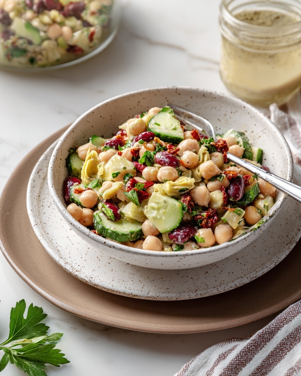 A white speckled bowl filled with a three-layer bean salad, featuring a mix of beige chickpeas, white cannellini beans, and dark red kidney beans scattered throughout. Slices of green cucumber, pieces of light yellow artichoke hearts, small bits of green leafy herbs, and sun-dried tomato fragments add pops of green, yellow, and dark red colors. A silver fork rests on the edge of the bowl, which sits on a plain white plate over a brown plate, all placed on a white marbled surface. In the background, a jar with a light dressing and a striped cloth napkin are partially visible. A green parsley leaf is on the white marbled surface in the foreground. Photo taken with an iphone --ar 4:5 --v 7