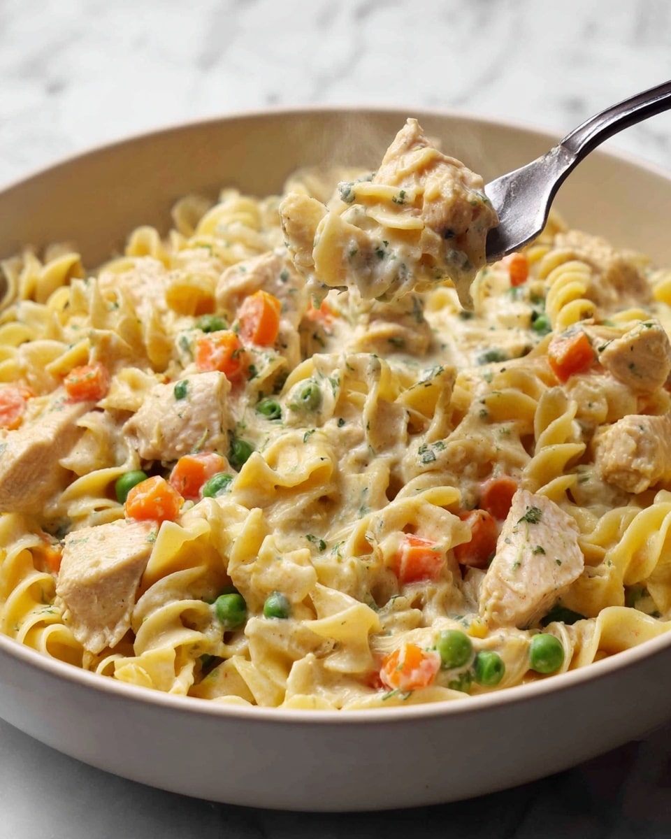 A white bowl filled with creamy pasta with three main layers: the base of curly egg noodles with a soft yellow color, scattered chunks of light brown chicken on top, and small diced bright orange carrots and green peas mixed evenly in the creamy sauce that coats everything with a pale off-white texture; steam rising gently from the dish suggests it is hot and fresh, and a silver fork holds a bite of pasta in the right side of the bowl, showing the thick sauce dripping slightly; the bowl sits on a white marbled surface. photo taken with an iphone --ar 4:5 --v 7