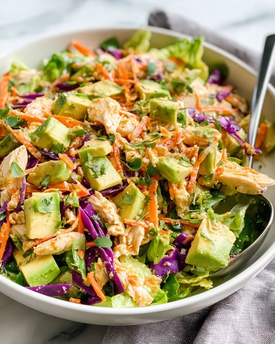 A close-up view of a fresh salad in a white bowl with a spoon inside. The salad has several layers including chunks of light green avocado, shredded purple cabbage, thin strips of orange carrot, and torn green lettuce leaves mixed evenly. Small pieces of light beige chicken are scattered throughout, adding texture. Sprinkled on top are tiny green herbs. The background is a white marbled texture with a light gray cloth visible under the bowl. photo taken with an iphone --ar 4:5 --v 7