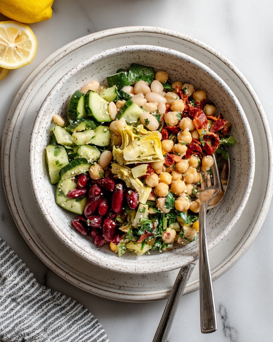 A white speckled bowl filled with a colorful salad made of multiple layers: creamy white cannellini beans, golden chickpeas, bright green cucumber slices, chopped spinach or herbs, deep red kidney beans, and small pieces of pale yellow artichoke hearts mixed with bits of sun-dried tomatoes. The textures vary from smooth beans to crisp cucumber and leafy greens. A silver fork rests inside the bowl, and the bowl sits on two stacked plain white plates. The surface beneath is white marble, with a striped cloth and a cut lemon nearby. Photo taken with an iphone --ar 4:5 --v 7