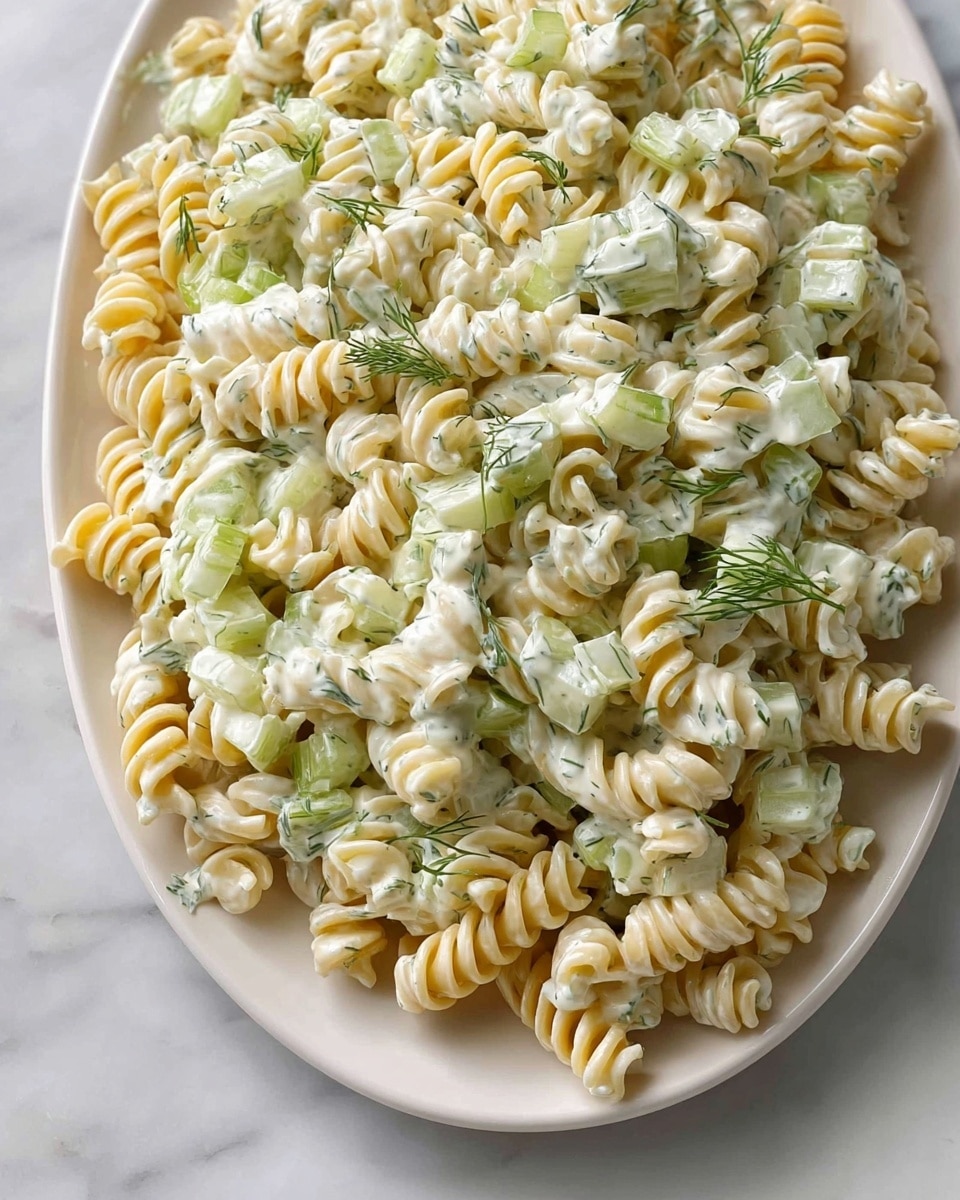 A white plate filled with creamy pasta salad, showing three main layers: the bottom layer is pale yellow rotini pasta with tight spirals; the middle layer includes light green, thinly sliced celery pieces spread throughout; the top layer is a thick white dressing with small green flecks of fresh dill sprinkled evenly, adding texture and color contrast. The whole dish looks fresh and mixed, sitting on a white marbled surface. photo taken with an iphone --ar 4:5 --v 7