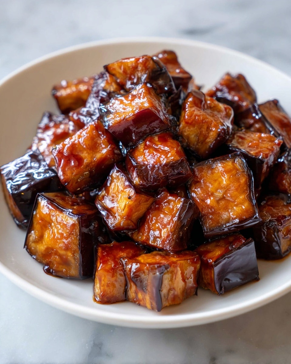A close-up view of a white plate filled with about three layers of shiny, dark brown glazed eggplant cubes. Each cube has a rich caramelized texture, the sauce thickly coating each piece and reflecting light. The eggplant skin appears deep purple, almost black, contrasting with the softer orange-brown interior visible on some sides. The plate sits on a white marbled surface that softly blurs in the background. Photo taken with an iphone --ar 4:5 --v 7