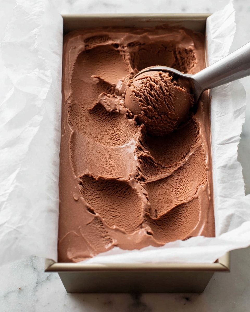 The image shows a close-up of a rectangular metal container filled with smooth, creamy chocolate ice cream. The ice cream has a rich dark brown color and a soft, dense texture, with a scoop taken out in the middle, showing the velvety inside. The scoop is held next to the container, with clear marks where the ice cream was cut, and the ice cream surface has gentle, uneven waves from being spread. The container is lined with white parchment paper that slightly wrinkles around the edges, all placed on a white marbled surface. photo taken with an iphone --ar 4:5 --v 7