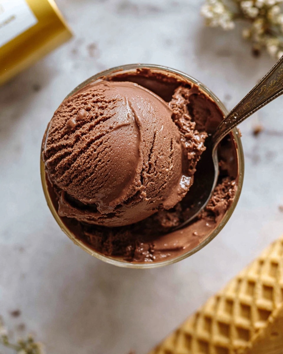 A close-up top view of a single, thick scoop of rich, creamy chocolate ice cream in a clear glass cup, showing its smooth and slightly glossy texture with detailed ridges and tiny air pockets, a vintage silver spoon partially dipped into the ice cream on the right side. The cup is placed on a white marbled surface with a stack of golden waffle cones visible on the right edge, and a blurred gold tube with a white label in the top left corner. The scene has warm natural lighting highlighting the ice cream’s creamy texture. photo taken with an iphone --ar 4:5 --v 7