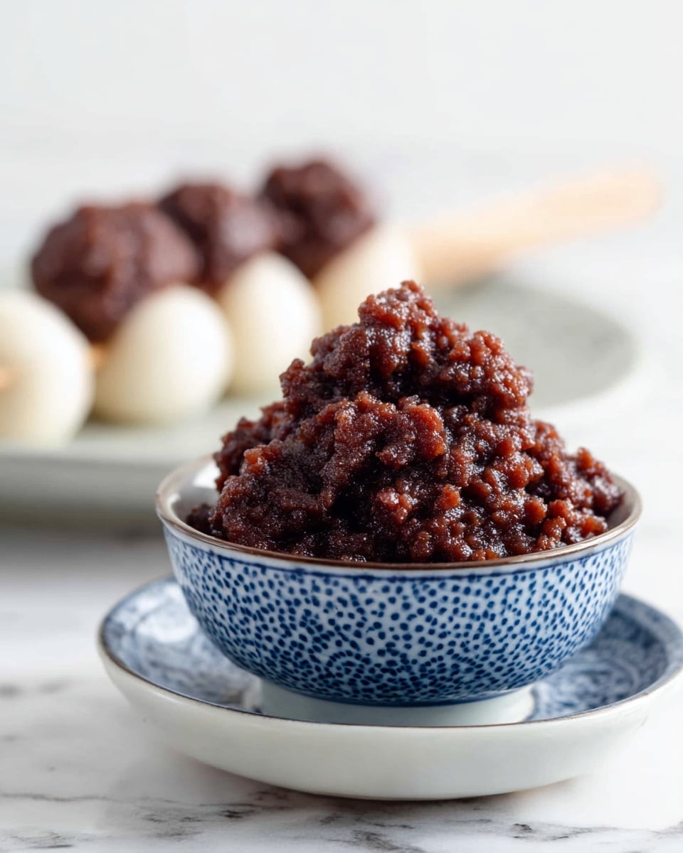 A close-up image shows a mound of chunky, dark reddish-brown sweet red bean paste piled in a blue and white patterned small bowl, which sits on a matching saucer. In the background, slightly out of focus, is a white plate holding skewered white rice dumplings topped with more red bean paste. The scene is set on a white marbled surface that gives a clean, fresh look. photo taken with an iphone --ar 4:5 --v 7