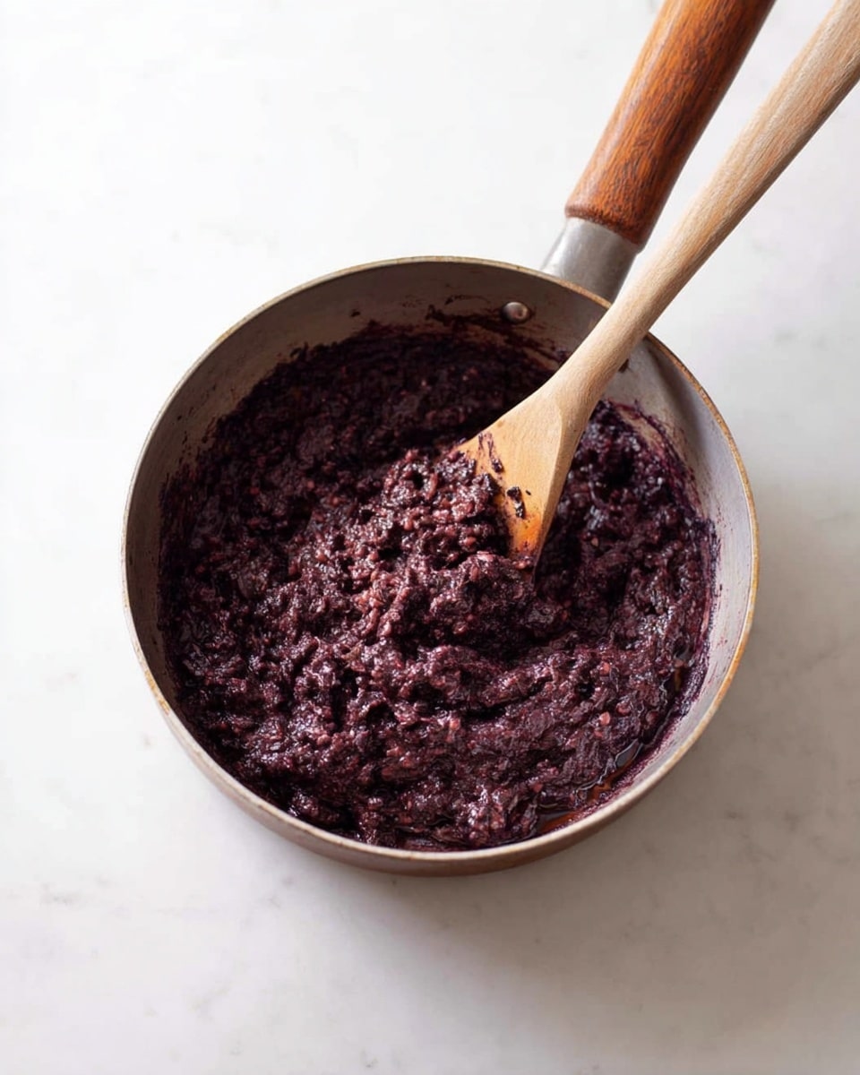 A metal pan with a wooden handle holds a thick, dark purple mixture with a chunky, mashed texture inside. A wooden spatula is partly submerged in the mixture, stirring it gently, showing some of the clumps and soft pieces clearly within the dense spread. The pan sits on a white marbled surface, and the lighting highlights the rich color and moist texture of the mixture inside the pan. photo taken with an iphone --ar 4:5 --v 7