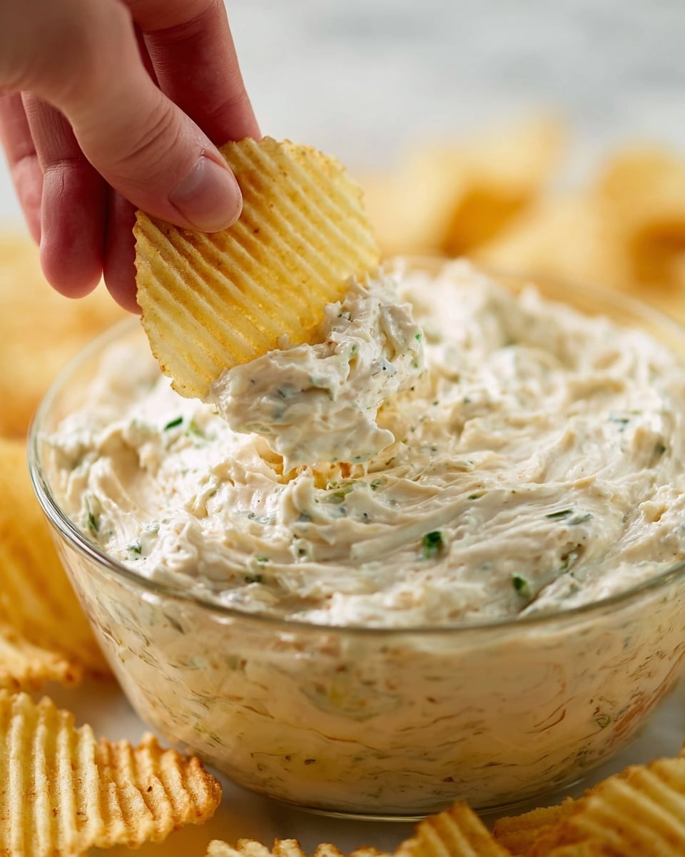 A close-up shows a clear glass bowl filled with a creamy dip that has a thick, chunky texture with small bits of green herbs mixed in. A woman's hand is dipping a ridged pale yellow potato chip into the dip, lifting some of it with the chip. In the background, more out-of-focus ridged potato chips are visible. The scene is set on a white marbled surface. photo taken with an iphone --ar 4:5 --v 7
