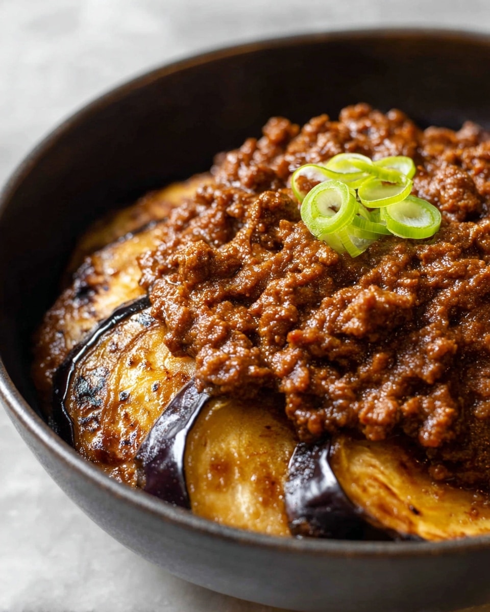 A close-up view of a dark bowl filled with two main layers: the bottom layer has several thick slices of golden brown roasted eggplant with visible charred edges, and the top layer is a generous mound of glossy, rich brown minced meat sauce with a saucy texture. On top of the meat, there are a few thin slices of bright green onion curls placed in the center. The bowl sits on a white marbled textured surface. photo taken with an iphone --ar 4:5 --v 7