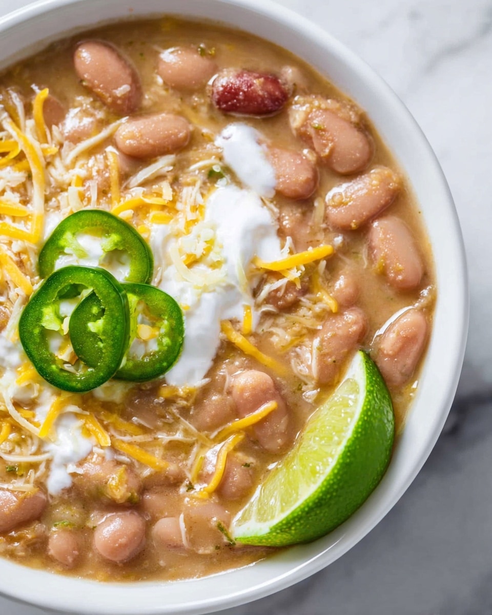 This close-up image shows a bowl filled with creamy, light brown beans mixed with whole pinkish beans and scattered shredded yellow cheese. On top, there are small dollops of white sour cream melting into the beans, a couple of thinly sliced green jalapeño pieces, and a wedge of bright green lime pressed into the side of the bowl. The bowl is white and the background is a white marbled texture. photo taken with an iphone --ar 4:5 --v 7