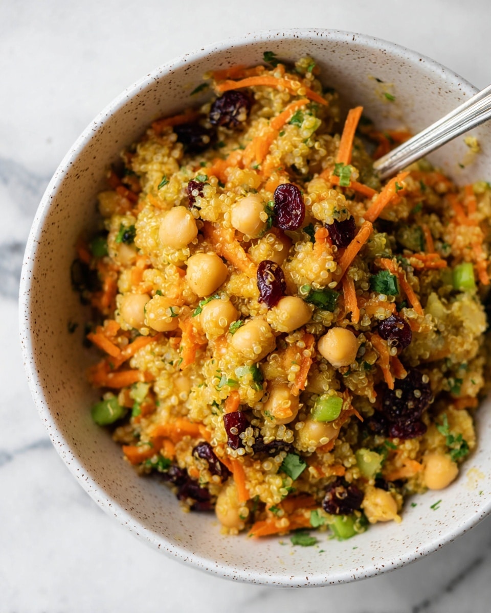 A close-up view of a bowl filled with a colorful quinoa salad showing about three layers of ingredients: the base layer is yellowish quinoa grains, mixed with light beige chickpeas, thin orange carrot shreds, small dark red dried cranberries, and green chopped herbs and celery pieces scattered throughout. The bowl is white with small dark speckles, and a silver spoon rests inside the bowl on the top right. The background is a white marbled texture. photo taken with an iphone --ar 4:5 --v 7