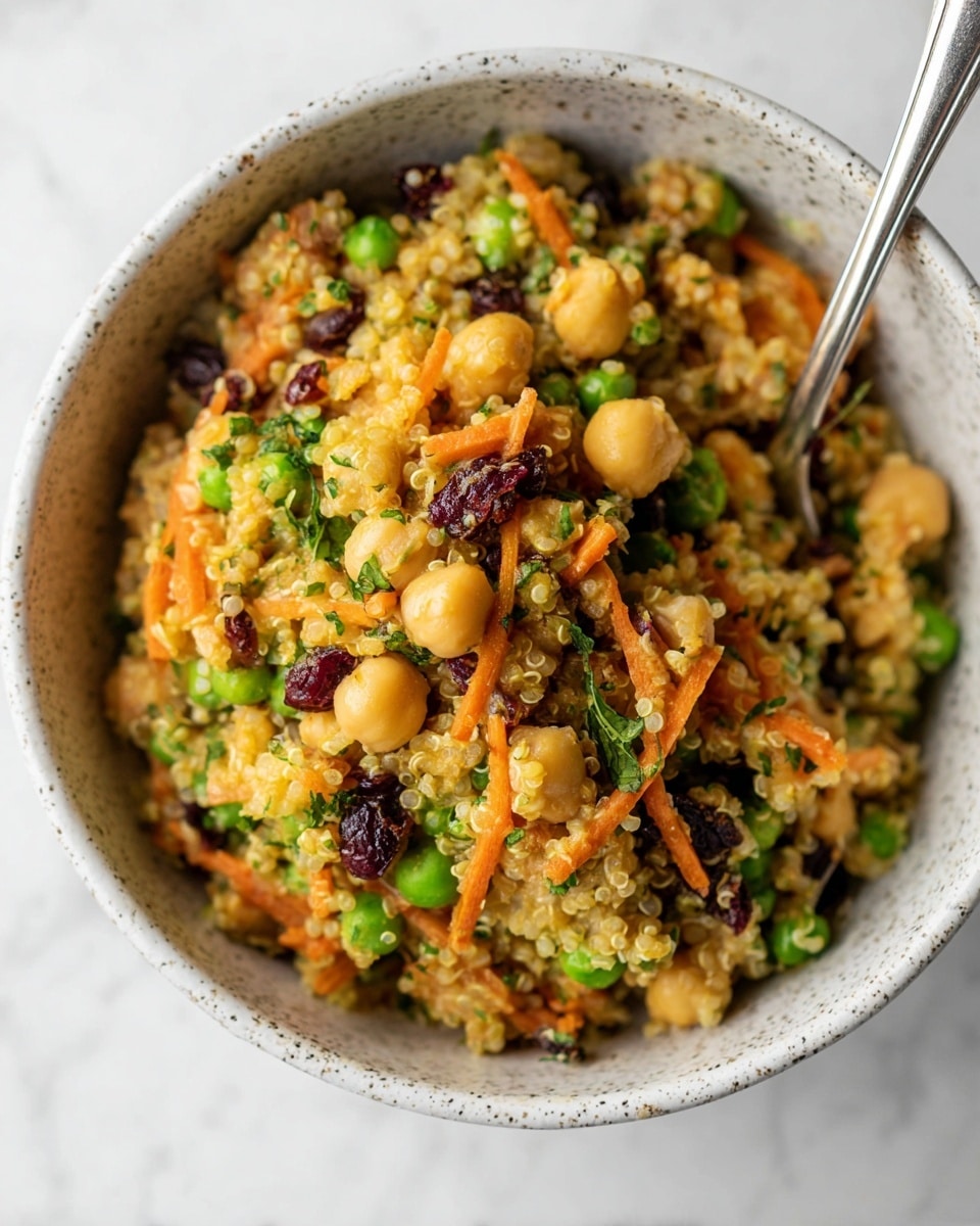A close-up view of a speckled white bowl filled with a colorful quinoa salad, showing a mix of beige chickpeas, light yellow quinoa grains, thin orange carrot shreds, small dark purple dried cranberries, and bright green chopped herbs and peas, with a silver spoon resting inside the bowl at the top right, all placed on a white marbled surface. photo taken with an iphone --ar 4:5 --v 7