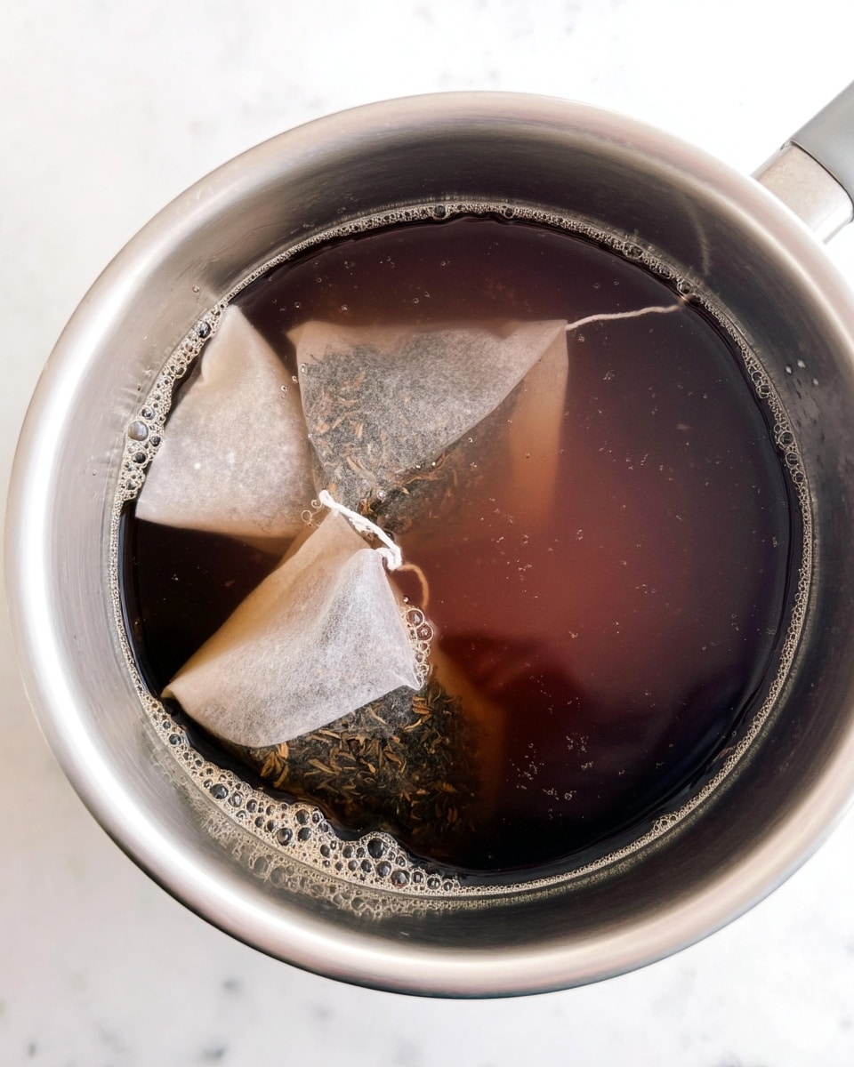 A close-up view inside a silver pot filled with dark brown tea liquid, showing three tea bags steeping. The tea bags are white and transparent, with visible tea leaves inside, floating near the pot's edge. The water around the tea bags has a light frothy layer, and the pot is placed on a white marbled surface. Photo taken with an iphone --ar 4:5 --v 7