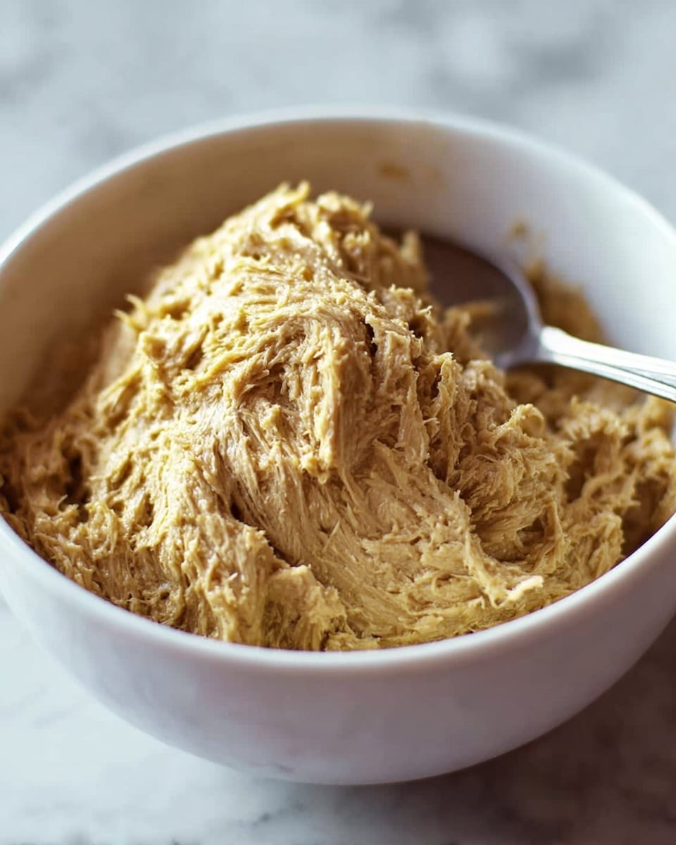 A close-up view of a white bowl filled with shredded, moist beige-yellow food that has a fibrous and textured appearance. The food forms a small peak in the center with a spoon partially inserted on the right side, creating a slight indentation. The background features a soft, white marbled texture, making the dish the clear focus. photo taken with an iphone --ar 4:5 --v 7
