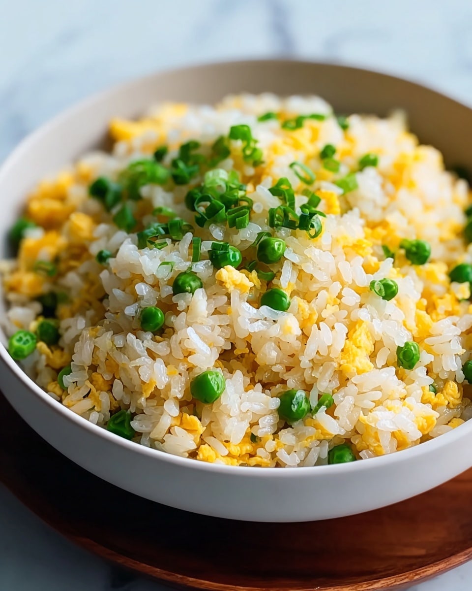 A close-up view of a white bowl filled with fried rice that has three main layers: fluffy white rice mixed with small yellow scrambled egg pieces, bright green chopped scallions scattered on top, and tiny green peas spread evenly throughout. The rice looks soft and slightly shiny, with the yellow eggs adding a textured look. The bowl is set on a wooden surface with a white marbled background softly blurred behind. Photo taken with an iphone --ar 4:5 --v 7
