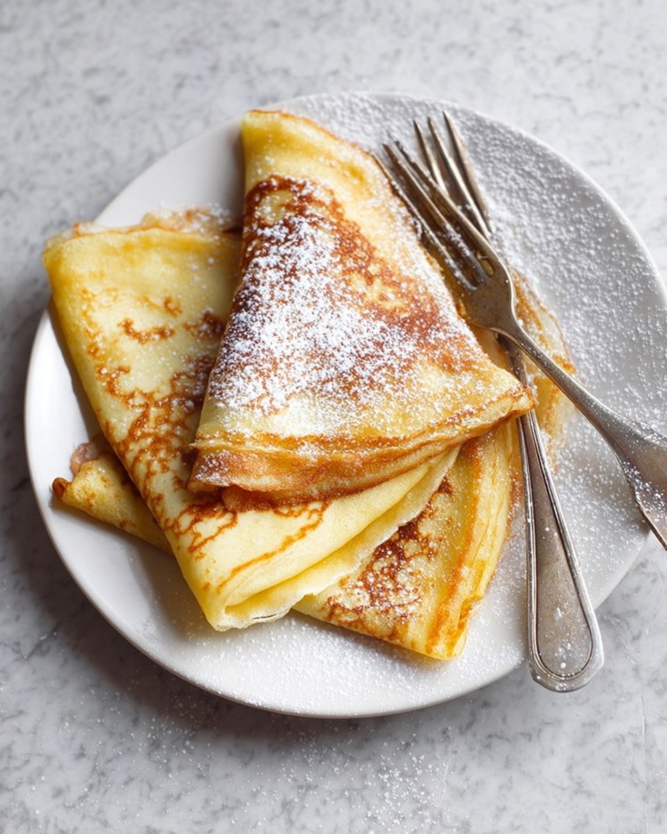 The image shows a white plate with three folded crepes, each folded into a quarter triangle shape. The crepes have a light golden yellow color with darker brown spots from cooking, giving them a soft, slightly crispy texture in some parts. A light dusting of white powdered sugar covers the crepes, adding a snowy texture on top. Two vintage silver forks rest on the plate, placed near the right edge. The plate sits on a white marbled surface with some scattered powdered sugar around it. Photo taken with an iphone --ar 4:5 --v 7