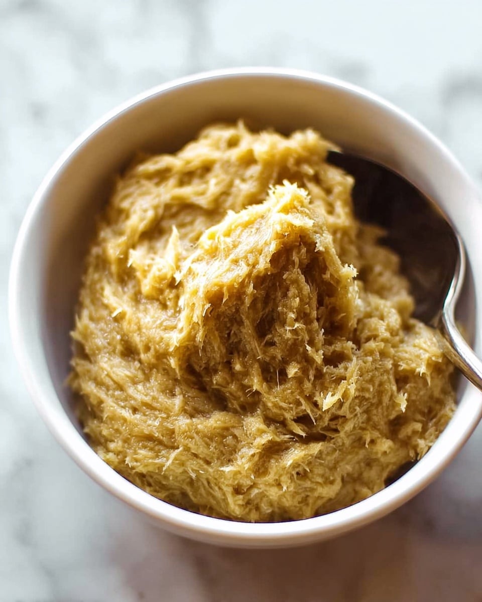 A close-up image of a creamy dish inside a white bowl, showing one thick, textured layer with a golden yellow color and fibrous bits visible throughout. The surface has a swirled, soft look as if stirred by a spoon, which is placed on the right side, partially visible. The bowl sits on a white marbled surface, giving a clean and bright background. Photo taken with an iphone --ar 4:5 --v 7