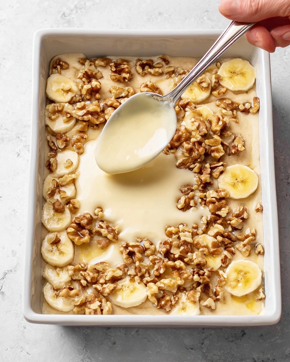 A white baking dish filled with a mixture layered with sliced banana pieces and small walnut chunks spread evenly on top. A creamy, light-colored batter or sauce is being spooned over the banana and walnut layer, creating small pools of smooth texture on the surface. The dish is set on a white marbled texture background, and a woman's hand is holding the spoon with batter. The overall appearance shows a mix of yellow, light brown, and cream colors with a rough texture from the nuts and softness from the banana slices. photo taken with an iphone --ar 4:5 --v 7