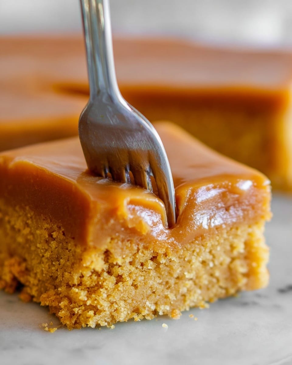 A close-up image of a two-layer dessert with a thick, smooth light brown caramel layer on top, and a dense, crumbly golden-brown cake layer beneath it. A silver fork is pressing gently into the top caramel layer, slightly breaking its smooth surface, showing the moist texture of the cake under the caramel. The background is a white marbled texture, creating a clean and soft contrast with the dessert. photo taken with an iphone --ar 4:5 --v 7