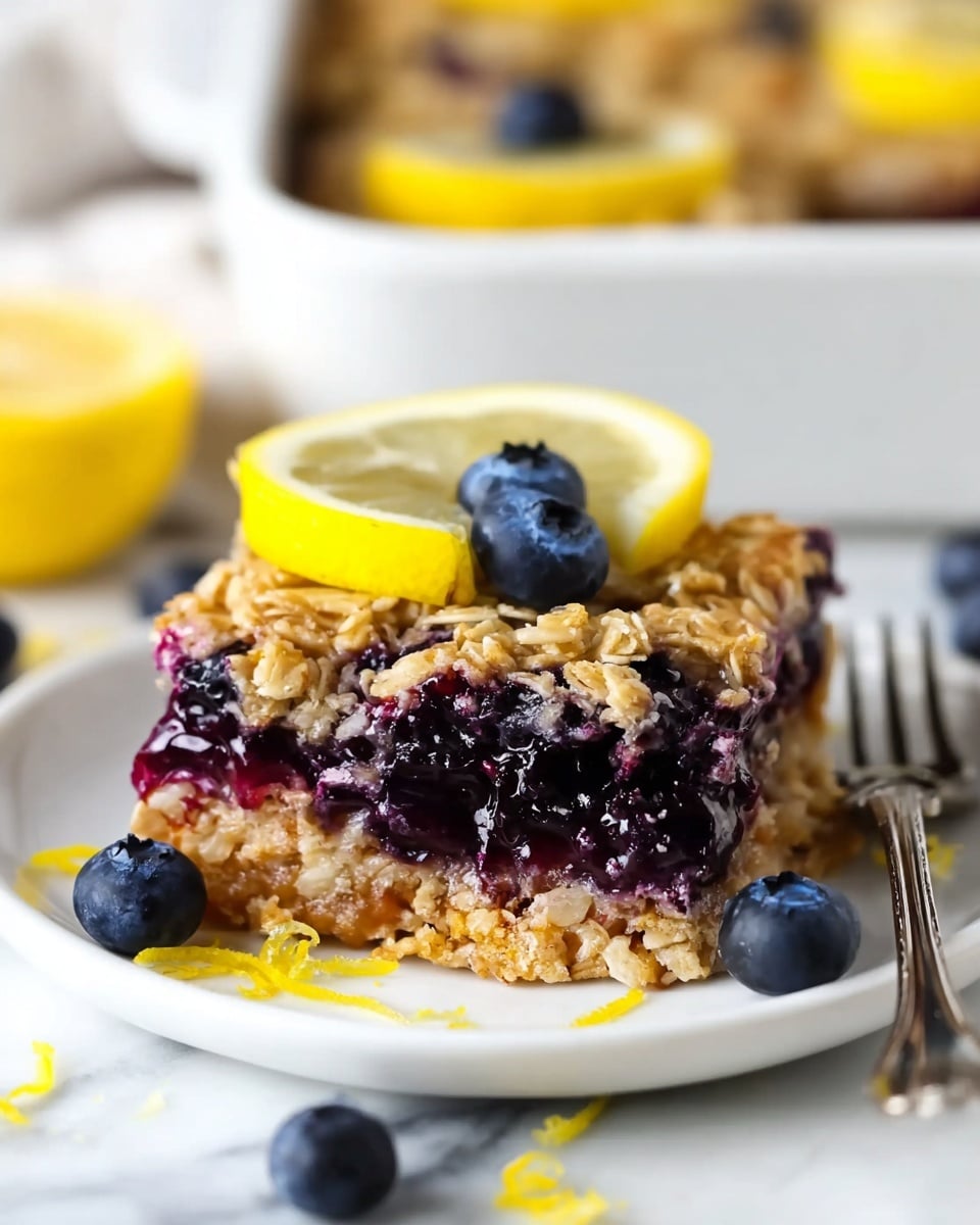 A close-up of a square blueberry oatmeal bar served on a white plate with a silver fork at the bottom right. The bar has two main layers: the top layer is a golden-brown oat crust with visible oats and a slightly coarse texture, and the bottom layer is filled with glossy, plump, dark purple blueberries mixed with oatmeal, creating a moist and juicy look. On top of the bar are two fresh blueberries and a thin bright yellow lemon slice, adding contrast. Scattered around the plate are a few fresh blueberries and small pieces of lemon zest. The background is a white marbled surface with a soft focus on a white baking dish filled with similar bars and lemon slices. Photo taken with an iphone --ar 4:5 --v 7