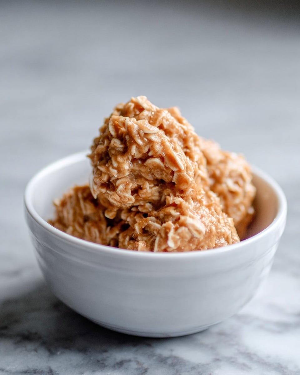 A white small bowl filled with three rough textured, chunky clusters of no-bake cookies, showing a mix of light brown and beige colors from peanut butter and oats. The cookies have uneven shapes and look soft and sticky. The bowl sits on a white marbled surface. Photo taken with an iphone --ar 4:5 --v 7