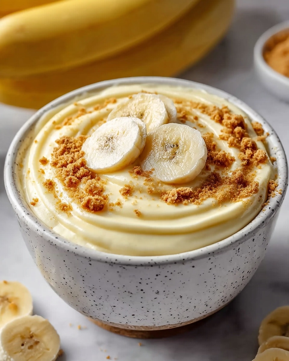 A white speckled bowl holds a creamy, smooth banana pudding with a swirled texture on top. The pudding is a pale yellow color, topped with crumbled light brown biscuit crumbs scattered over the surface. Three neatly arranged round banana slices rest in the center, adding a fresh touch. The bowl sits on a white marbled surface with blurred bananas in the background. Photo taken with an iphone --ar 4:5 --v 7
