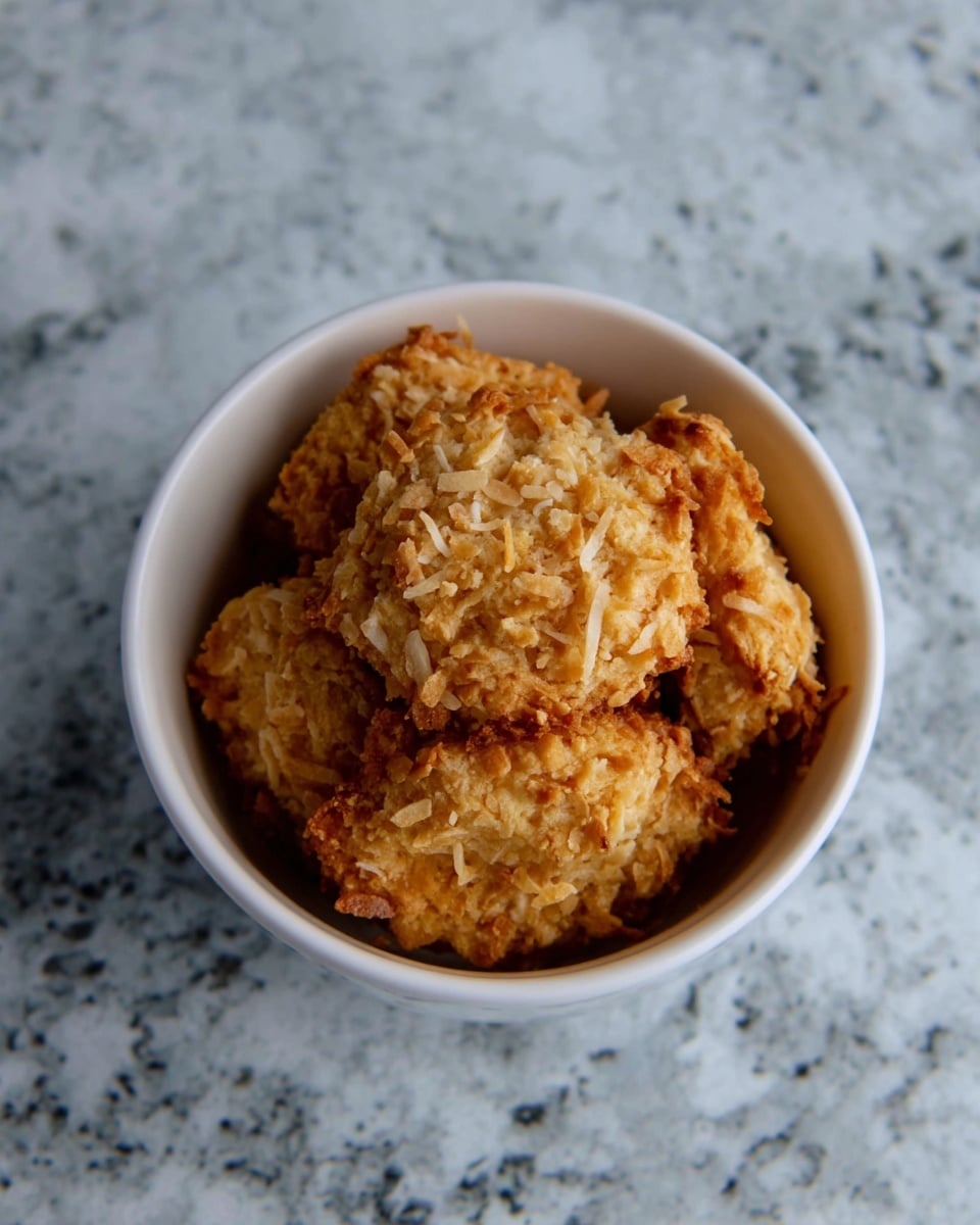A white bowl filled with several golden brown cookies that have a rough texture and visible shredded coconut on top, placed on a surface with a white marbled texture. photo taken with an iphone --ar 4:5 --v 7