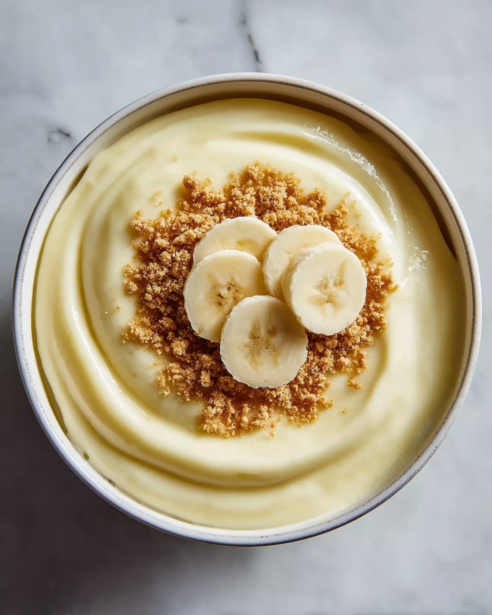 A white bowl holds a creamy light yellow pudding with a smooth, swirled texture filling the bowl. On top, there is a sprinkling of light brown crumbly crumbs forming a circular patch near the center. Three round, pale yellow banana slices are placed neatly on the crumbs, arranged in a small cluster. The bowl is set on a white marbled surface, showing slight reflections and soft natural light. photo taken with an iphone --ar 4:5 --v 7